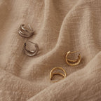 The Jenna Triple Hoops, minimalist silver and gold earrings, are shown on beige textured fabric—perfect for achieving a modern earring look.