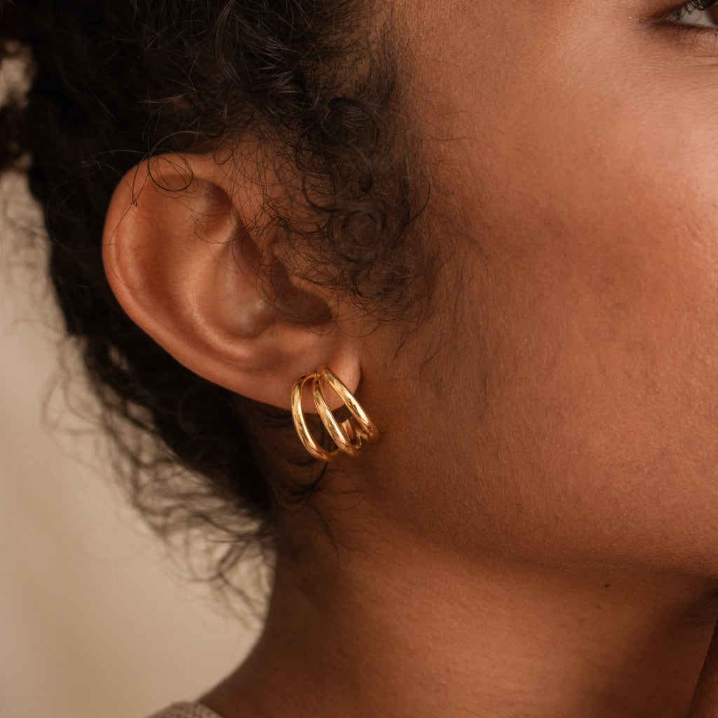 Close-up of a woman's ear wearing Jenna Triple Hoops, minimalist gold earrings, against a neutral background, highlighting their modern style.