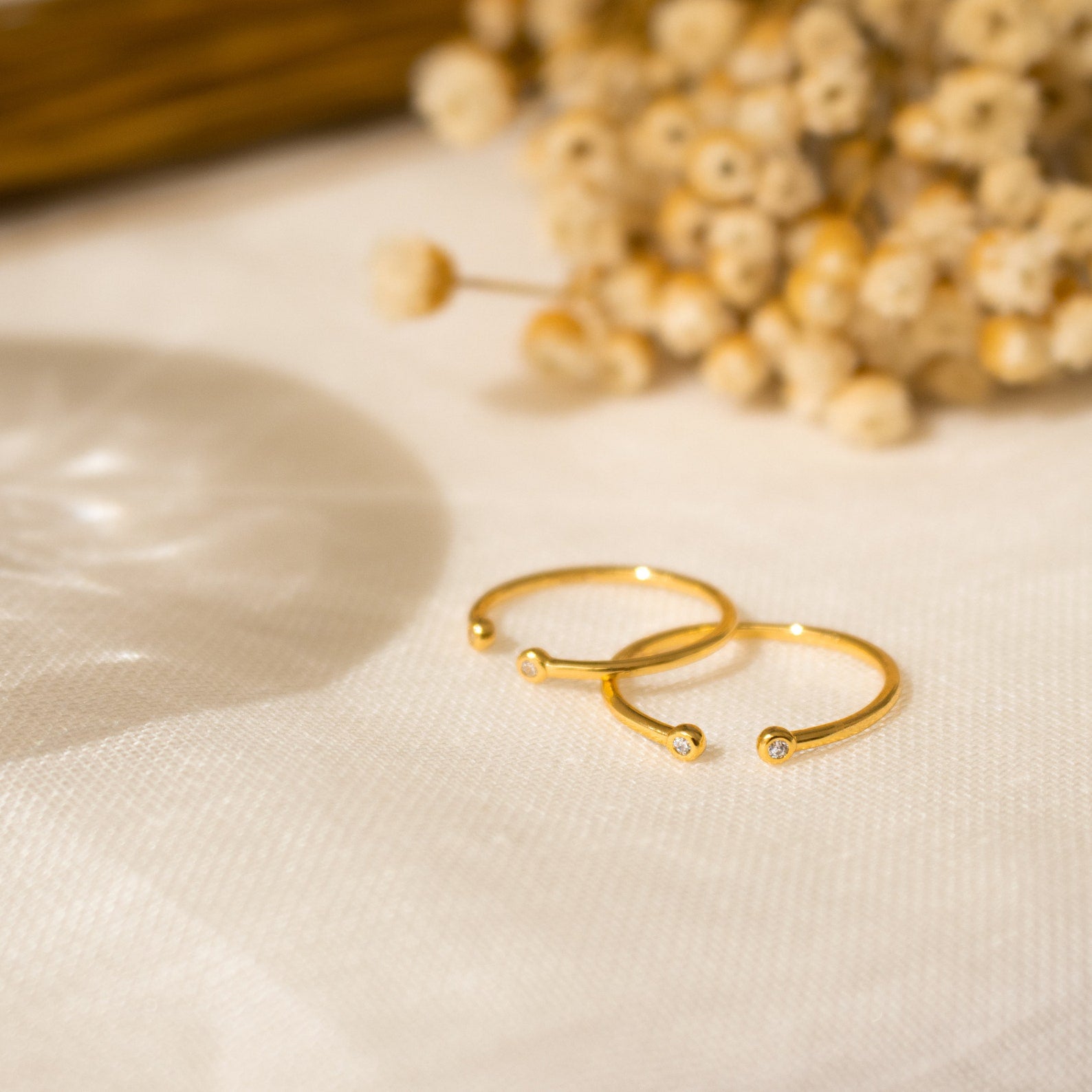 The adjustable Open Diamond Duo Ring in delicate gold rests on light fabric, with softly blurred dried flowers in the background—a perfect showcase of minimalistic jewelry.