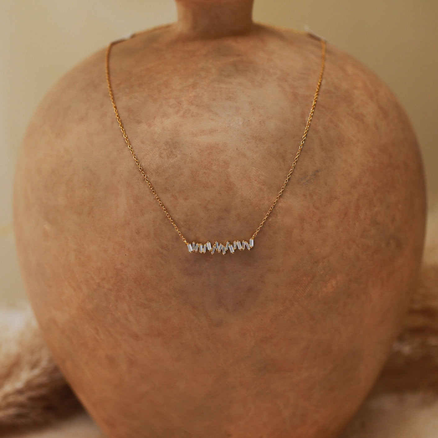 Gold necklace displayed on a terracotta surface, featuring a row of stacked, organic-shaped white beads arranged in a horizontal line at the center.