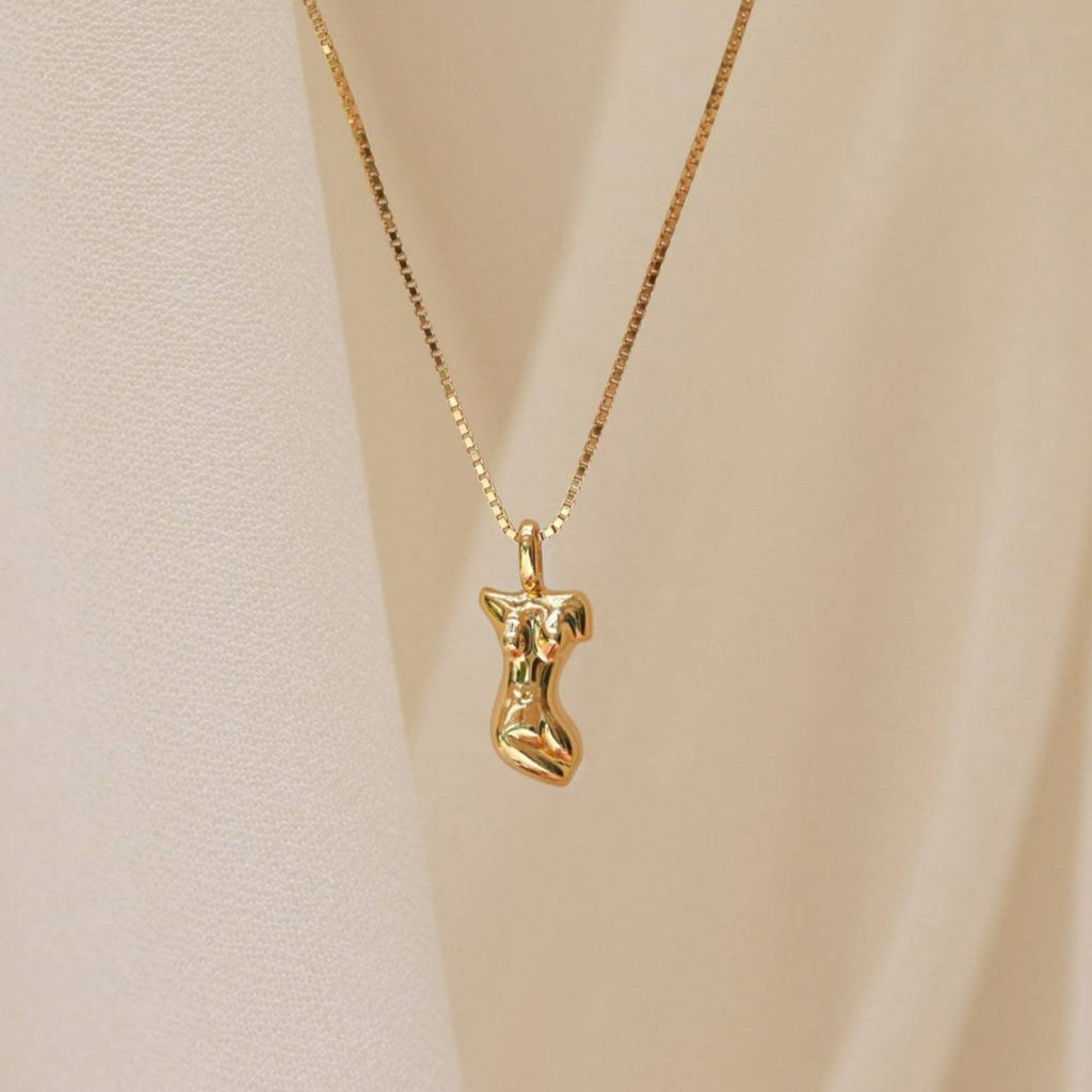 The Kinn Body Necklace features a gold pendant shaped like an abstract female torso, inspired by Venus de Milo, shown against a soft beige background.