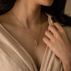 A woman wears the Kinn Body Necklace—a gold piece with a small pendant—resting near her collarbone, beautifully accenting her beige wrap top and showcasing empowerment jewelry that elevates her style.