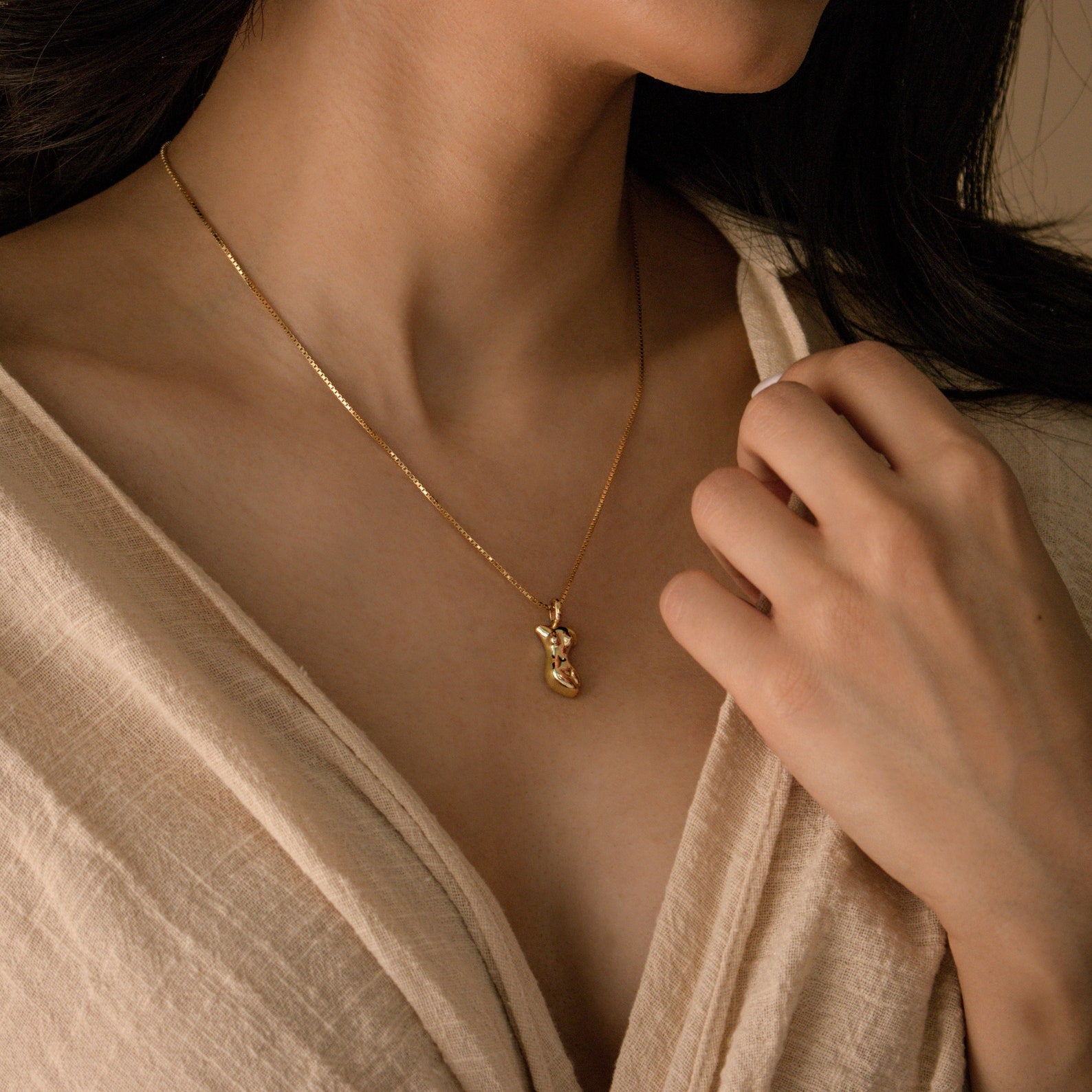 A woman wears the Kinn Body Necklace—a gold piece with a small pendant—resting near her collarbone, beautifully accenting her beige wrap top and showcasing empowerment jewelry that elevates her style.