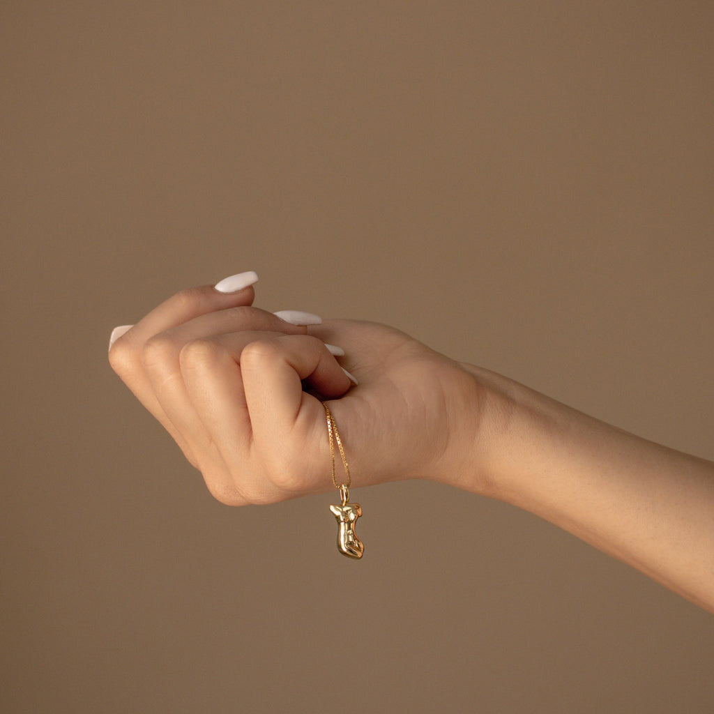 A hand with manicured nails holds the Kinn Body Necklace, featuring a gold boot-shaped pendant that symbolizes empowerment, set against a beige background.
