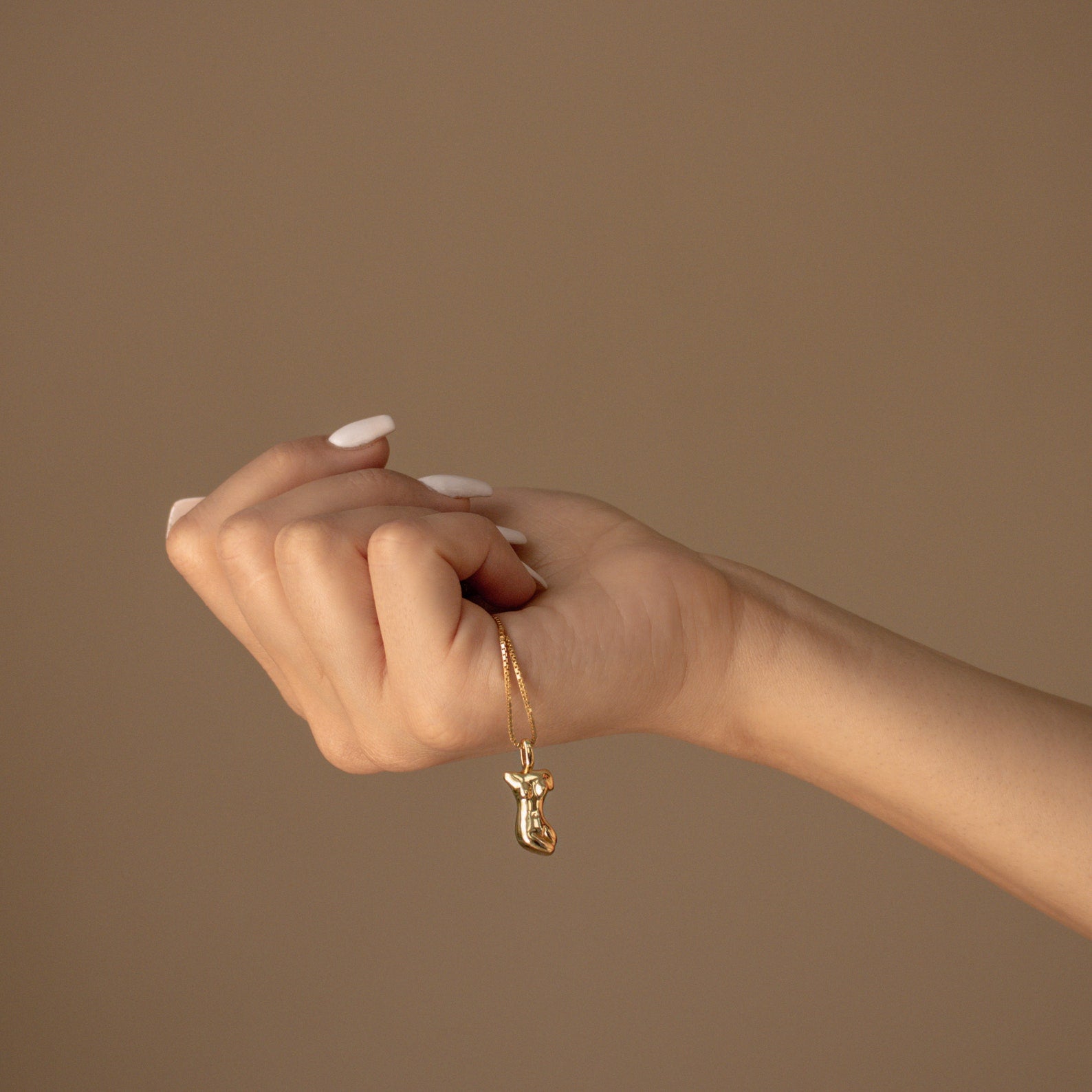 A hand with manicured nails holds the Kinn Body Necklace, featuring a gold boot-shaped pendant that symbolizes empowerment, set against a beige background.