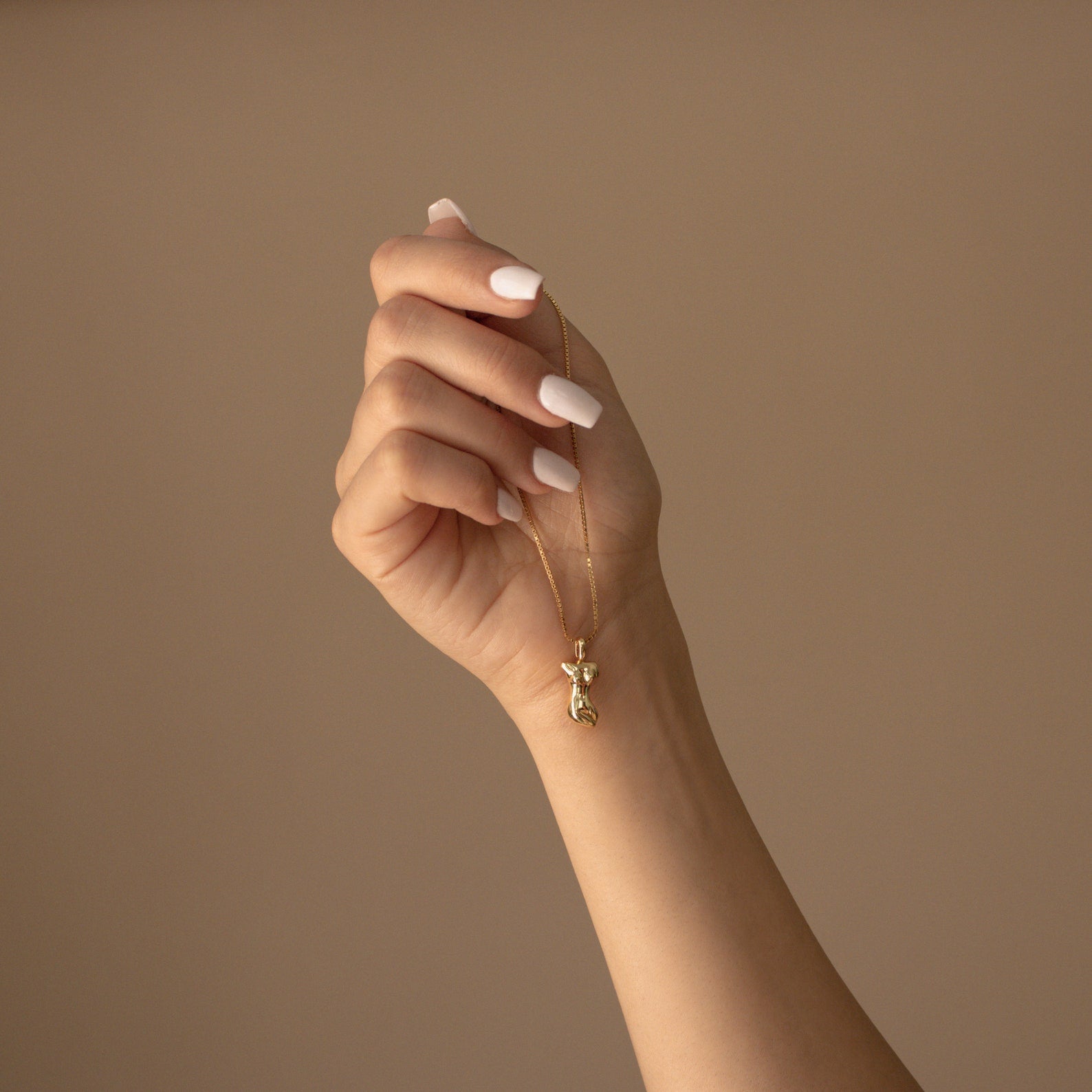 A manicured hand holds the Kinn Body Necklace, a gold Venus de Milo pendant symbolizing empowerment, against a beige background.
