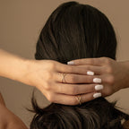 A woman with white manicured nails and gold rings, including the sparkling Pave X Ring set with tiny diamonds, holds the back of her head, her hair tied in a low bun.