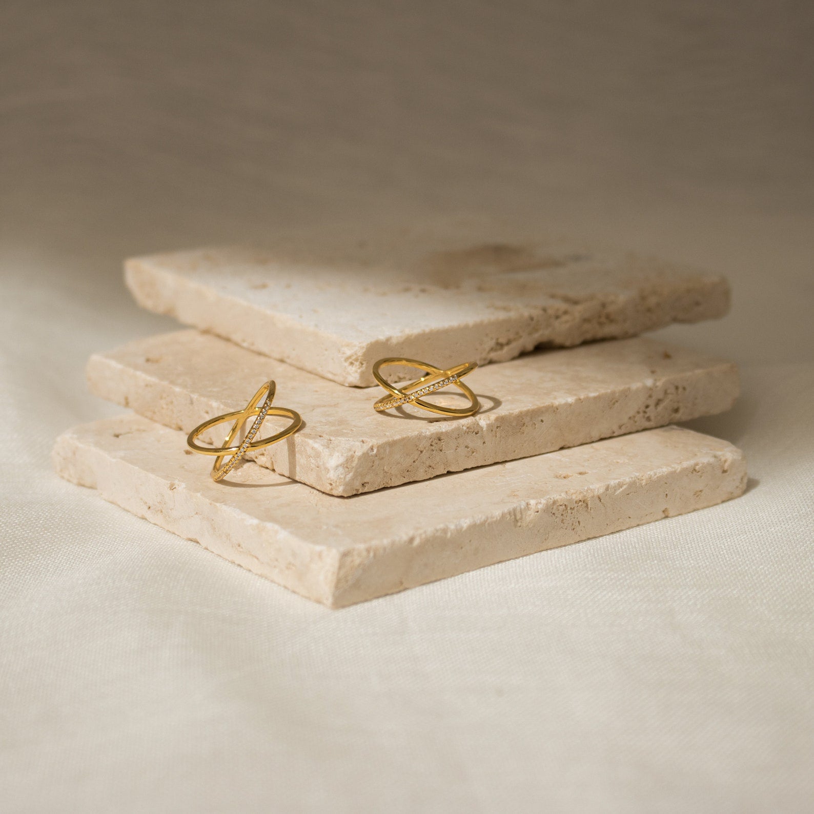 The Pave X Ring, a gold ring with intersecting bands and a pavé of tiny diamonds, is displayed on stacked stone tiles against a neutral background.