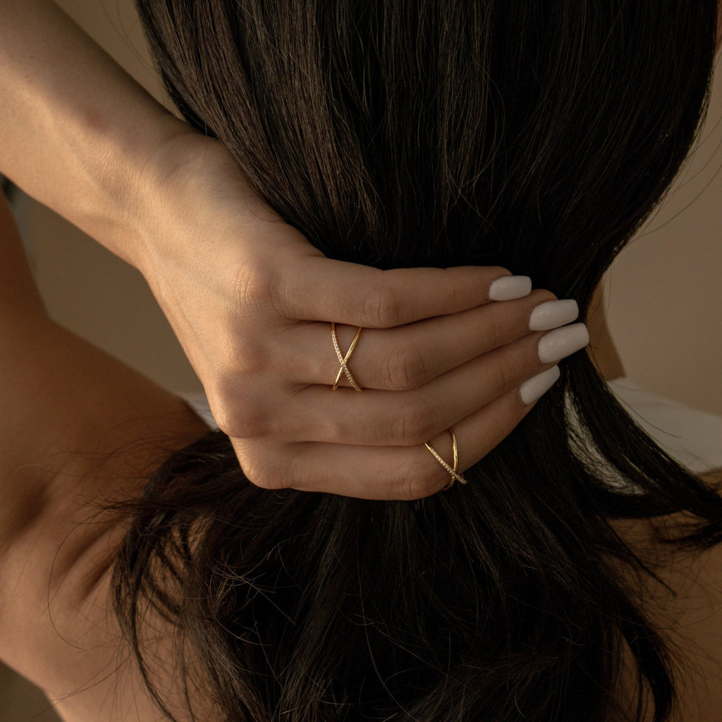 Hands with white nail polish hold dark hair, wearing two gold rings—one is the Pave X Ring sparkling with tiny diamonds.