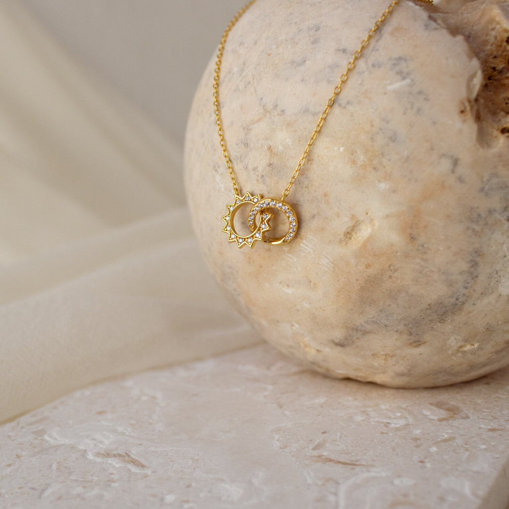 The Sun & Moon Eternity Necklace, elegantly displayed on a light stone with a neutral fabric background, beautifully captures the enchanting allure of both day and night.