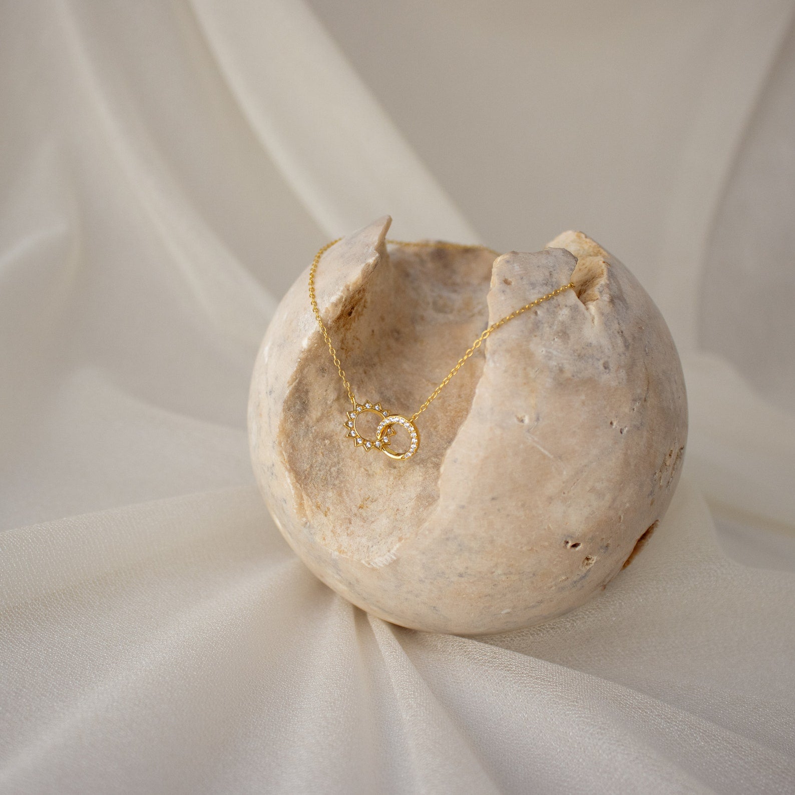 The Sun & Moon Eternity Necklace, featuring interlocking gold circles, rests gracefully on a round textured stone atop white fabric.