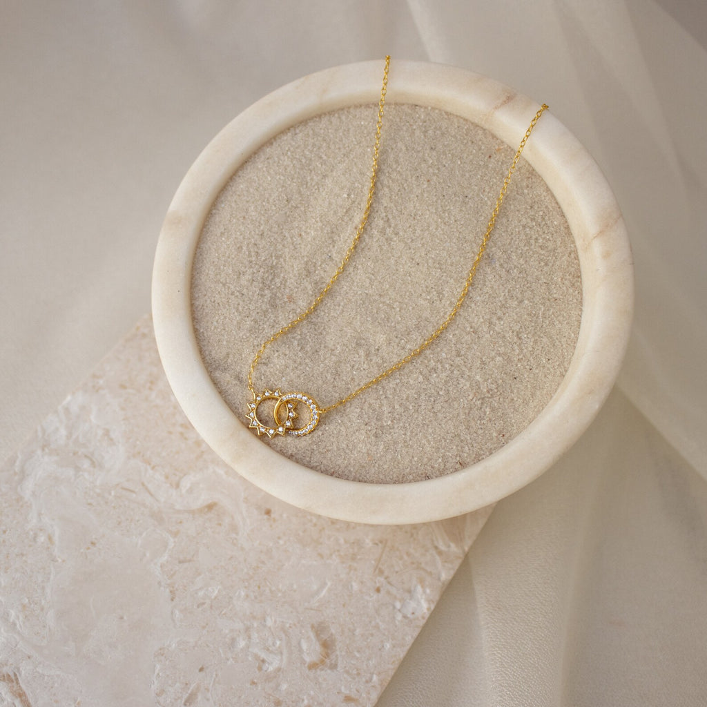 The Sun & Moon Eternity Necklace, with its interlocked circle pendants, is displayed on sand in a round marble dish, adding a celestial touch of elegance.