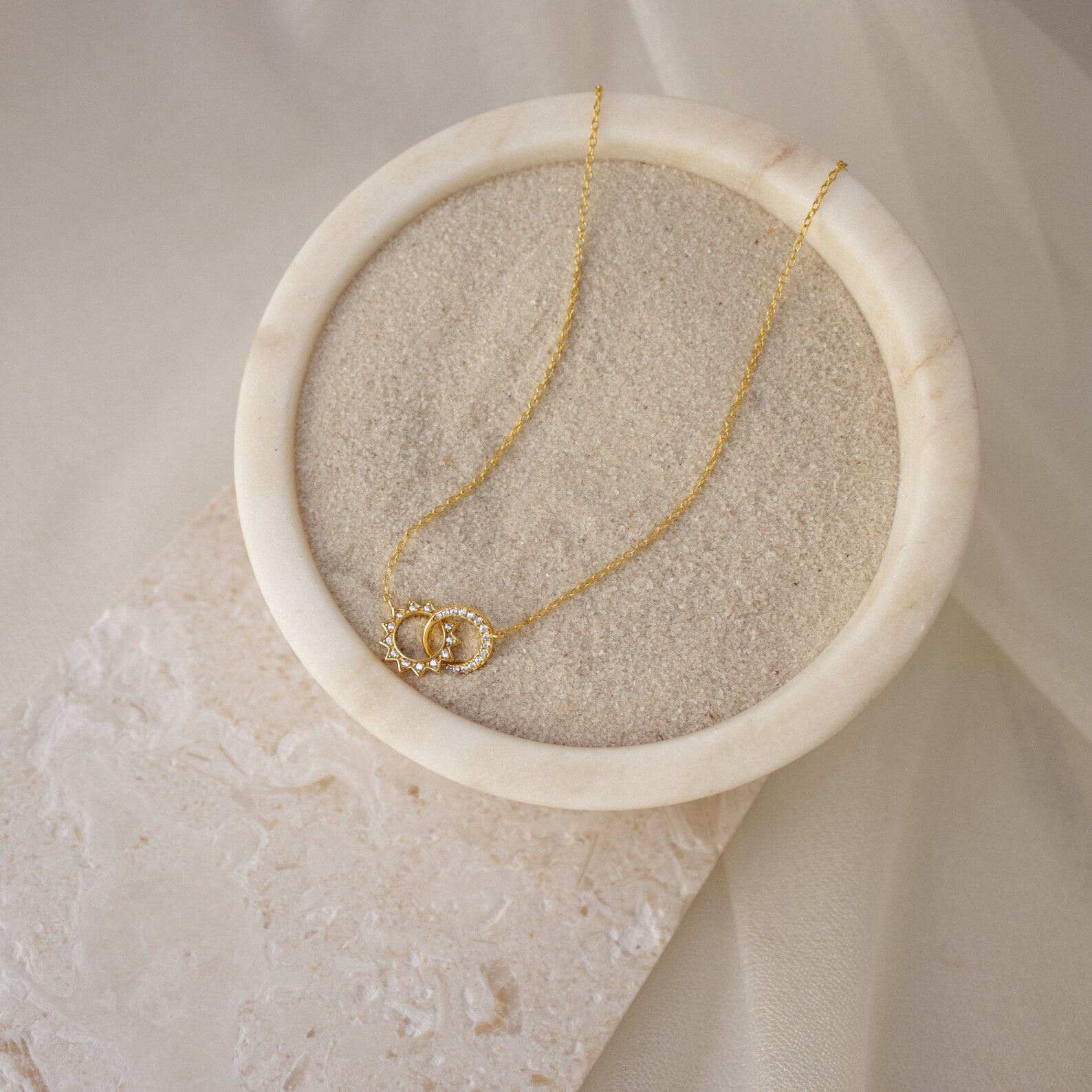 The Sun & Moon Eternity Necklace, with its interlocked circle pendants, is displayed on sand in a round marble dish, adding a celestial touch of elegance.