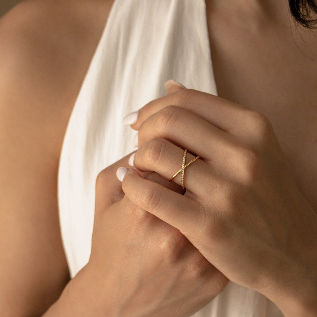 A person wearing a white halter top displays the Pave X Ring—a delicate gold ring adorned with sparkling pavé diamonds—on their finger, complemented by neatly manicured nails.