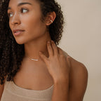 Woman with curly hair wears a beige top, gold ear cuff, and the Bethany Baguette Bar Necklace, looking to the side.