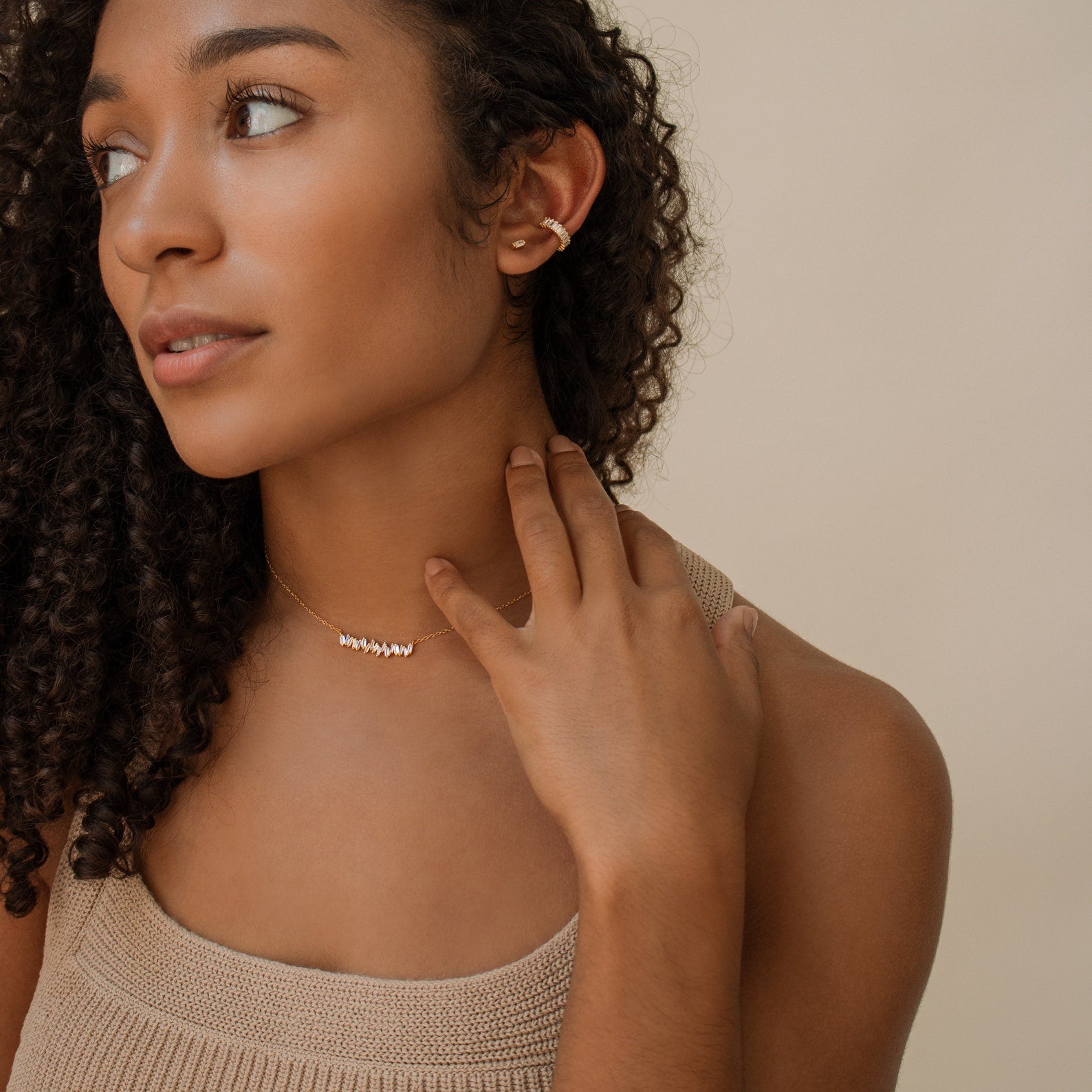 Woman with curly hair wears a beige top, gold ear cuff, and the Bethany Baguette Bar Necklace, looking to the side.