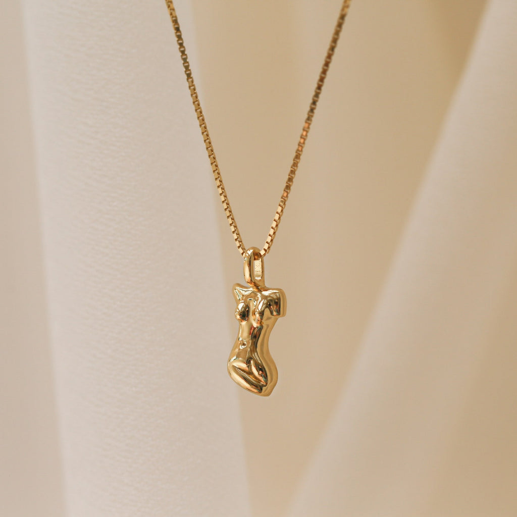 The Kinn Body Necklace features a gold pendant shaped like a stylized human torso, inspired by the Venus de Milo, displayed against a soft beige background—a refined and empowering jewelry piece.