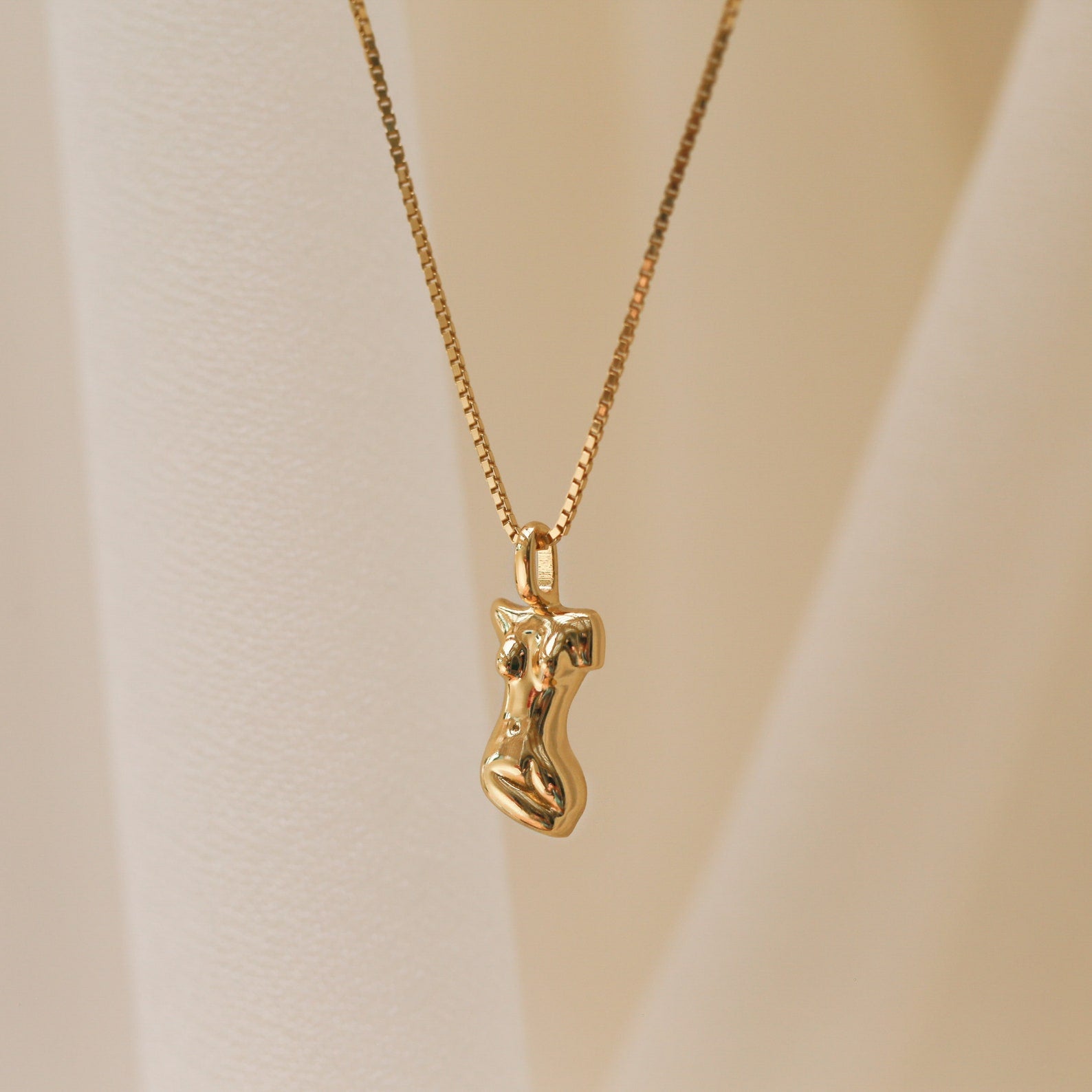 The Kinn Body Necklace features a gold pendant shaped like a stylized human torso, inspired by the Venus de Milo, displayed against a soft beige background—a refined and empowering jewelry piece.