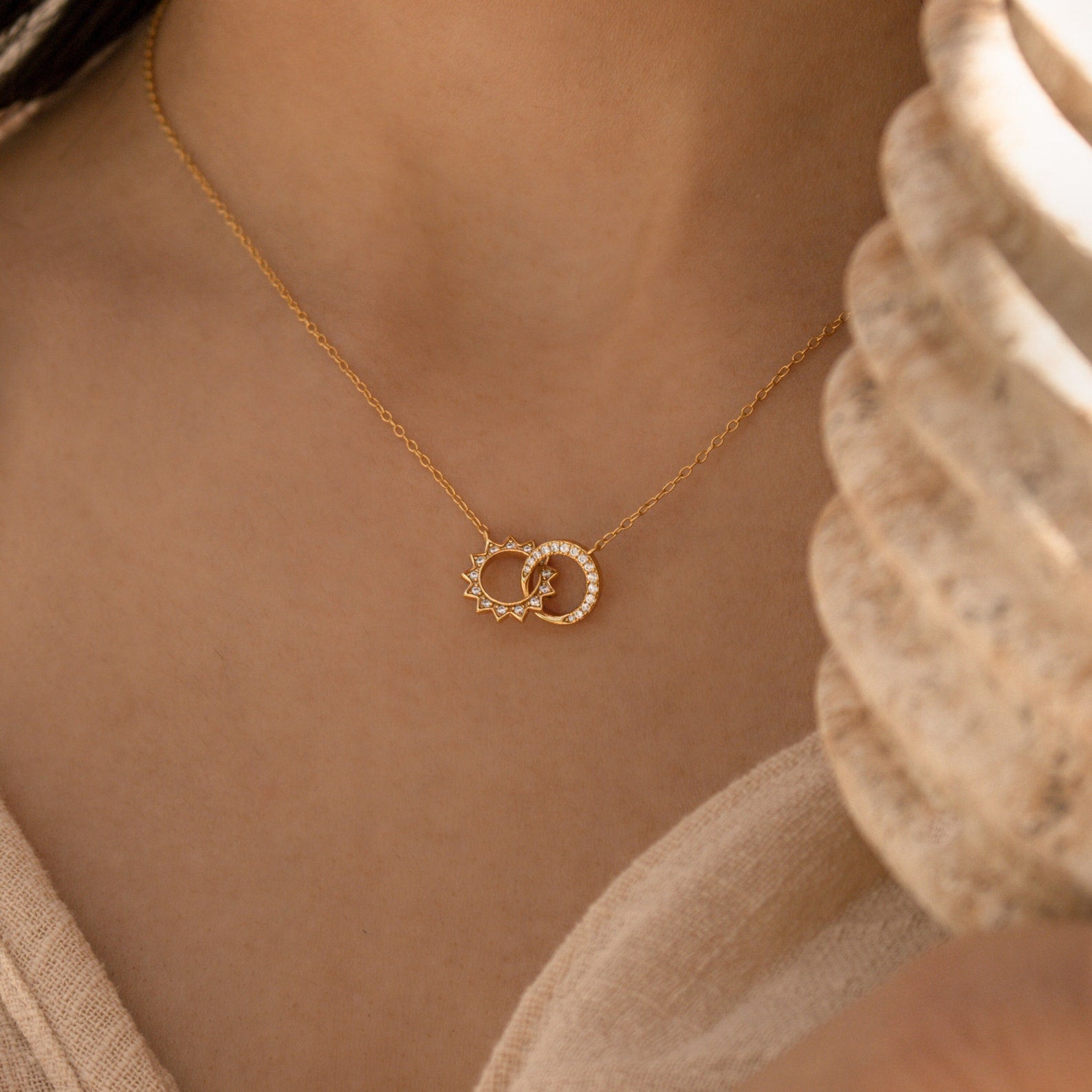 Close up of a woman wearing a gold Sun & Moon Eternity Necklace, featuring two interlocking circular charms—one shaped like a sunburst and the other pave-set with crystals—styled against a soft beige textured top.