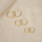 The Medium Thin Hoops earrings are shown with other gold hoop styles in different sizes on a light, textured fabric background.