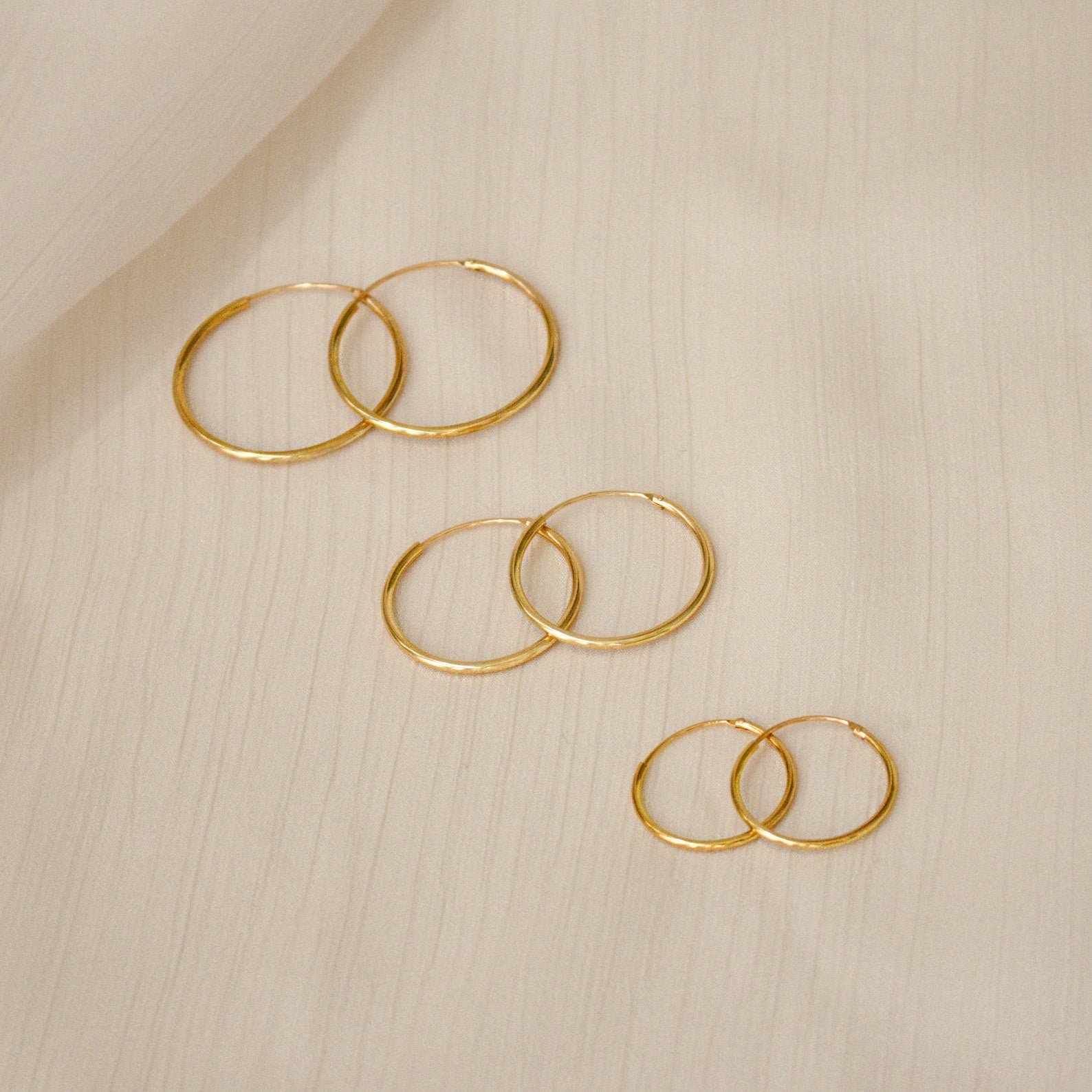The Medium Thin Hoops earrings are shown with other gold hoop styles in different sizes on a light, textured fabric background.