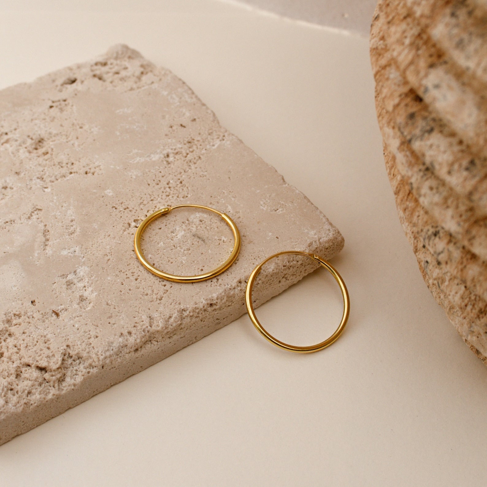 The Endless Thin Hoops, displayed on a textured beige stone beside a rough round accent, are timeless gold earrings ideal for daily wear.