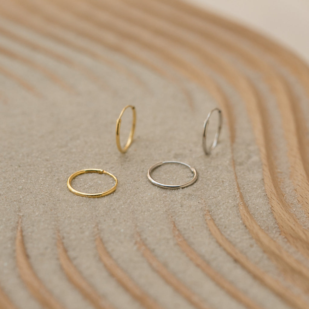 Two gold and two silver Small Thin Hoops rest on textured sand with curved wooden lines, perfect for elevating your everyday wardrobe.