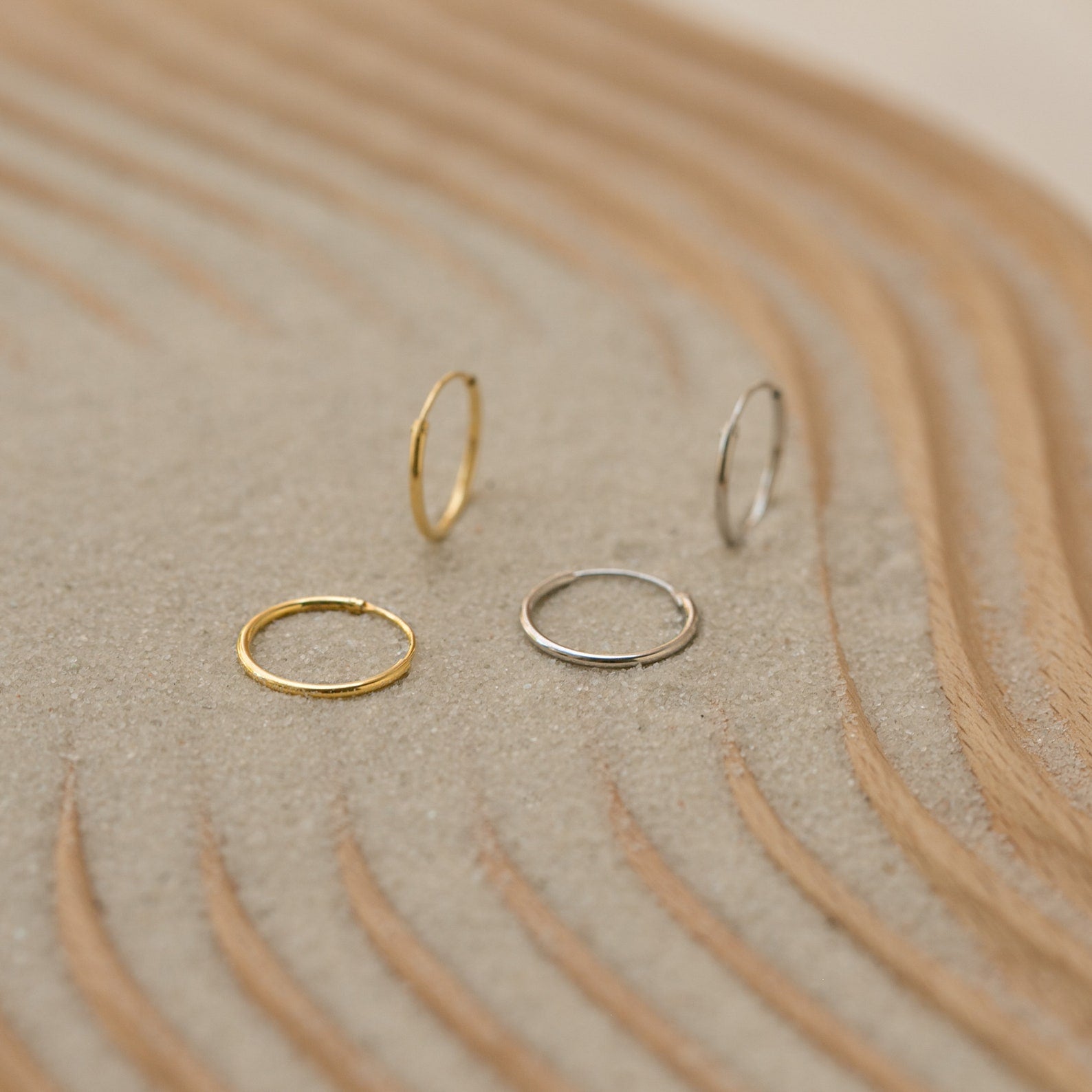Two gold and two silver Small Thin Hoops rest on textured sand with curved wooden lines, perfect for elevating your everyday wardrobe.