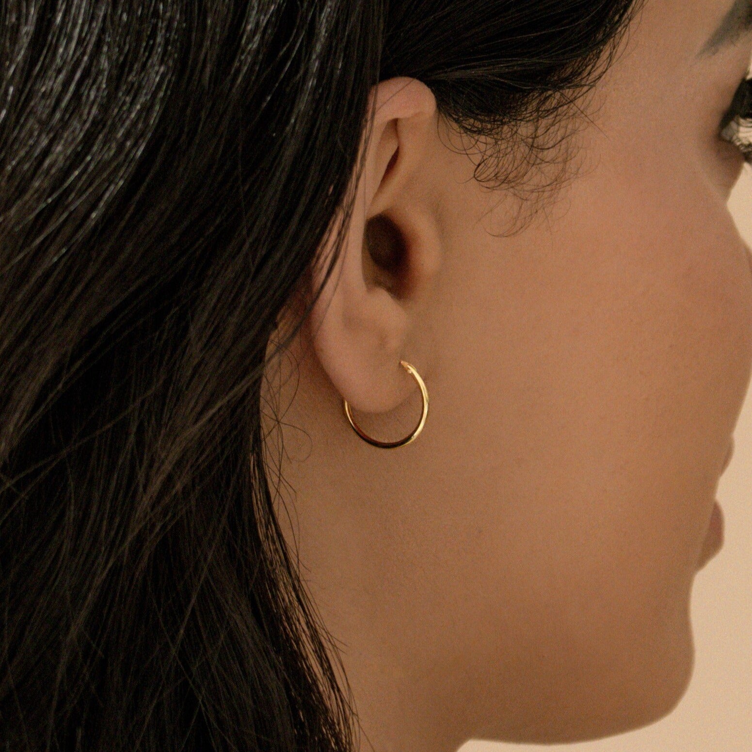 A close-up of a woman's ear showcases the Small Thin Hoops, her dark hair tucked behind her ear—an effortless accent for any everyday wardrobe.