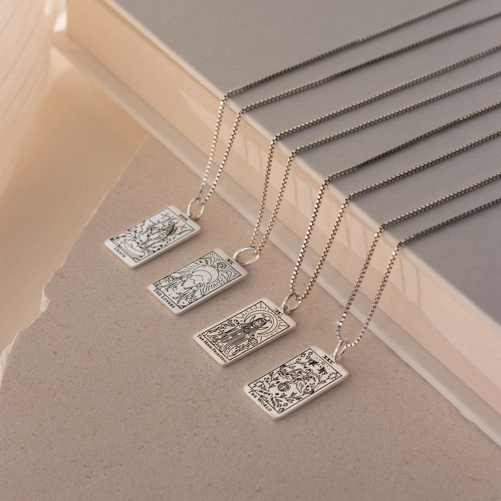 Four Tarot Necklace in Box Chain styles featuring rectangular Major Arcana pendants are displayed on a beige textured surface near books, perfect for adding mystique to your Tarot collection.