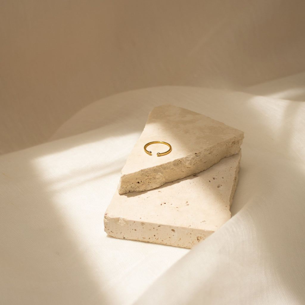 The Open Diamond Duo Ring, a minimalistic jewelry piece, is displayed on two beige stone slabs, highlighted by gentle sunlight and delicate shadows.