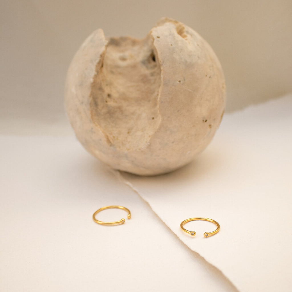 The Open Diamond Duo Ring, a minimalistic gold piece from our collection, is displayed on a cream surface with a textured, cracked sphere in the background.