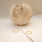 The Open Diamond Duo Ring, a minimalistic gold piece from our collection, is displayed on a cream surface with a textured, cracked sphere in the background.