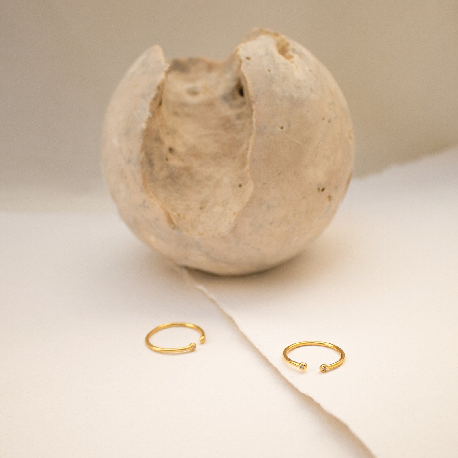 The Open Diamond Duo Ring, a minimalistic gold piece from our collection, is displayed on a cream surface with a textured, cracked sphere in the background.