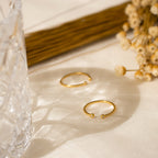 The Open Diamond Duo Ring, a delicate gold piece, rests on cream fabric next to dried flowers and cut glass in gentle sunlight.