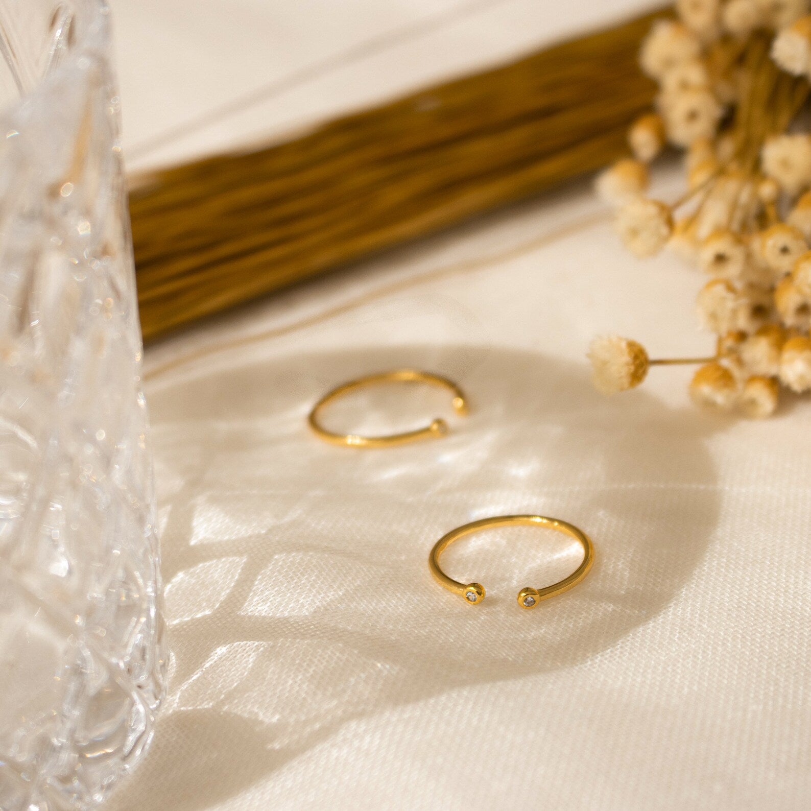 The Open Diamond Duo Ring, a delicate gold piece, rests on cream fabric next to dried flowers and cut glass in gentle sunlight.