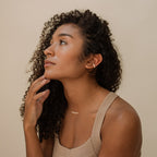 A woman with curly hair wears gold jewelry, including the Bethany Baguette Bar Necklace, and a beige top, looking upward against a neutral background.