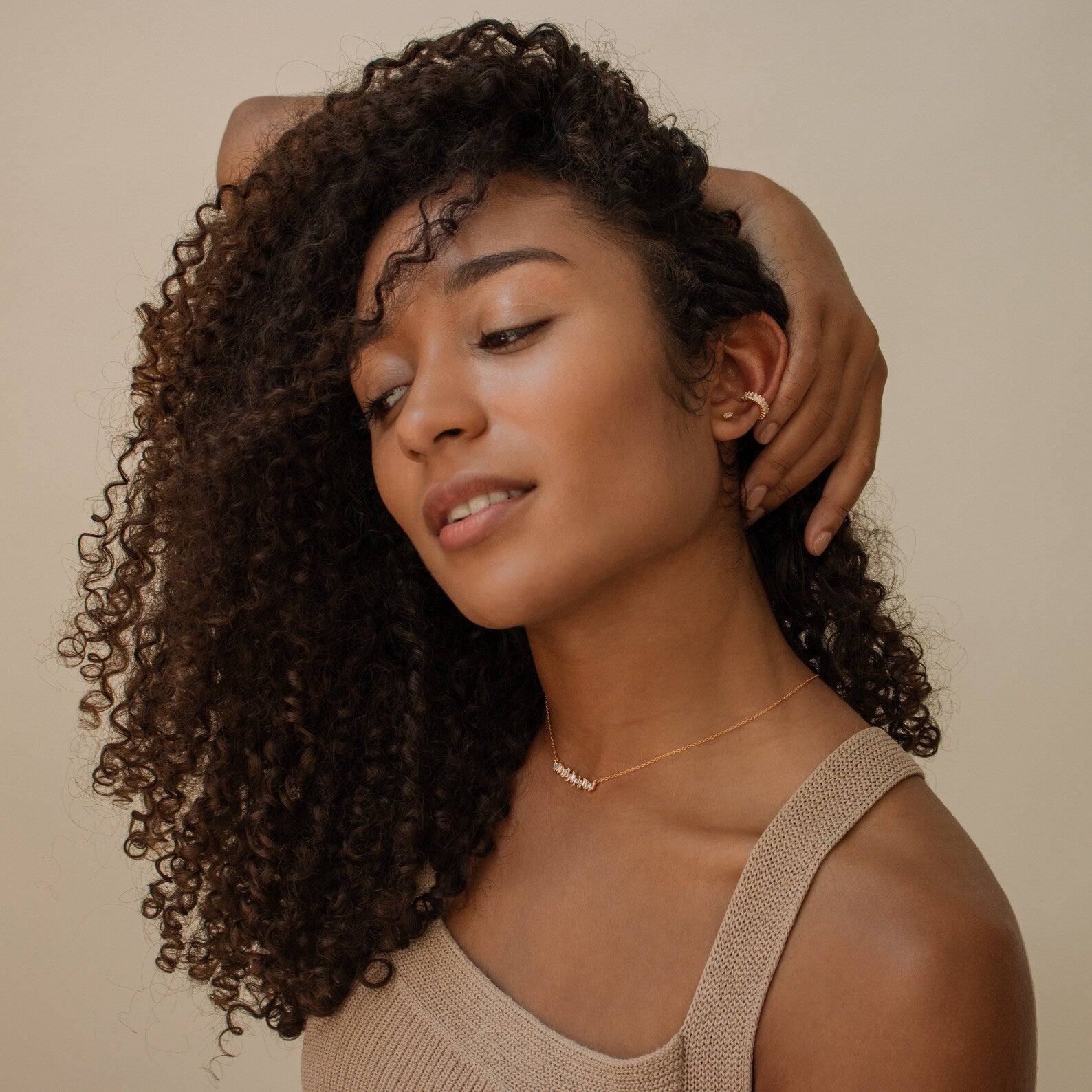 Woman with curly hair in a beige top, tilting her head and touching her ear, wearing the Bethany Baguette Bar Necklace—a delicate cable chain style.