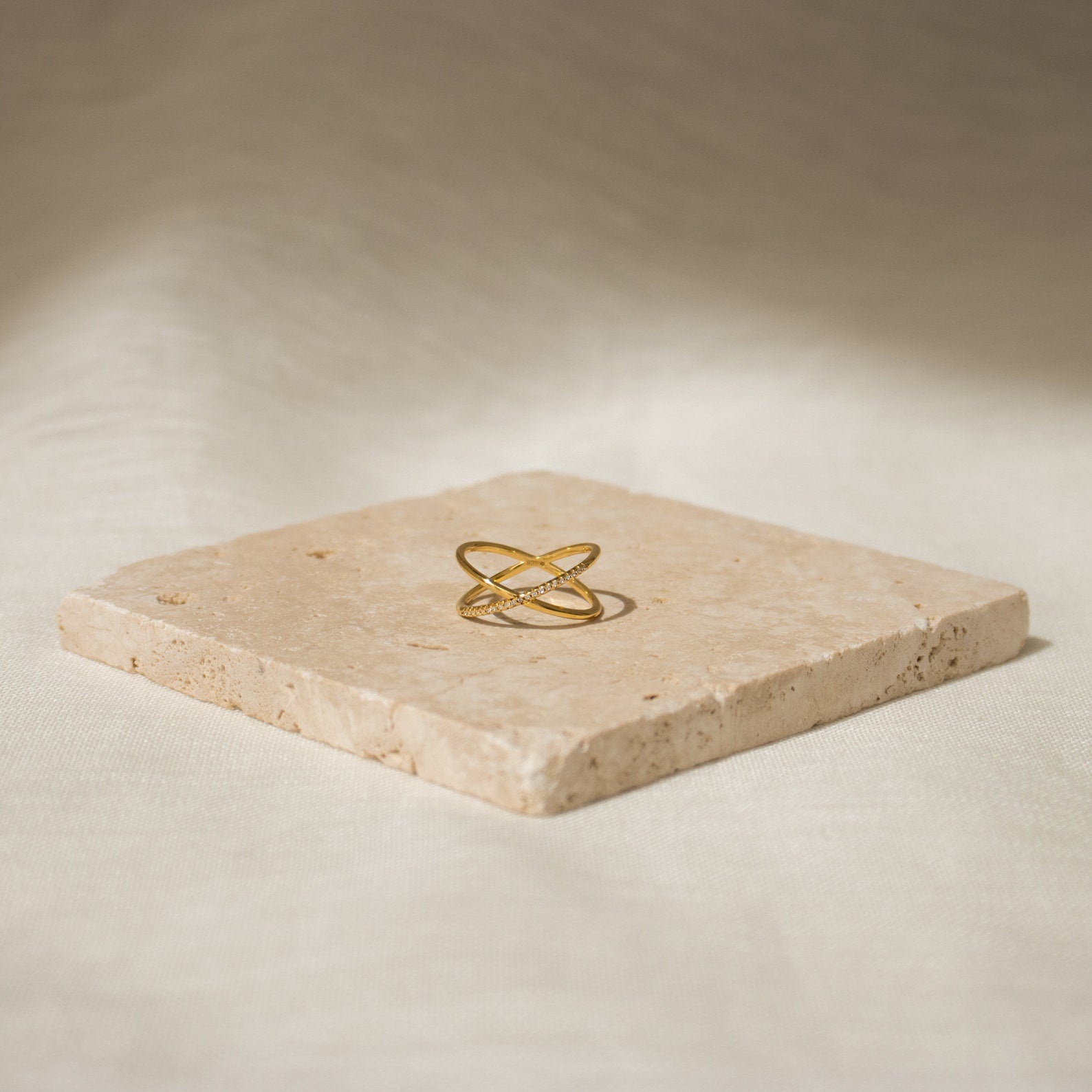 The Pave X Ring features a gold intersecting design with pavé diamonds, displayed on a square stone slab set against a neutral background.