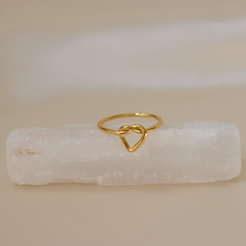 The Love Heart Knot Ring sits gracefully on a white rectangular crystal, making it an ideal promise ring or a timeless symbol of love.