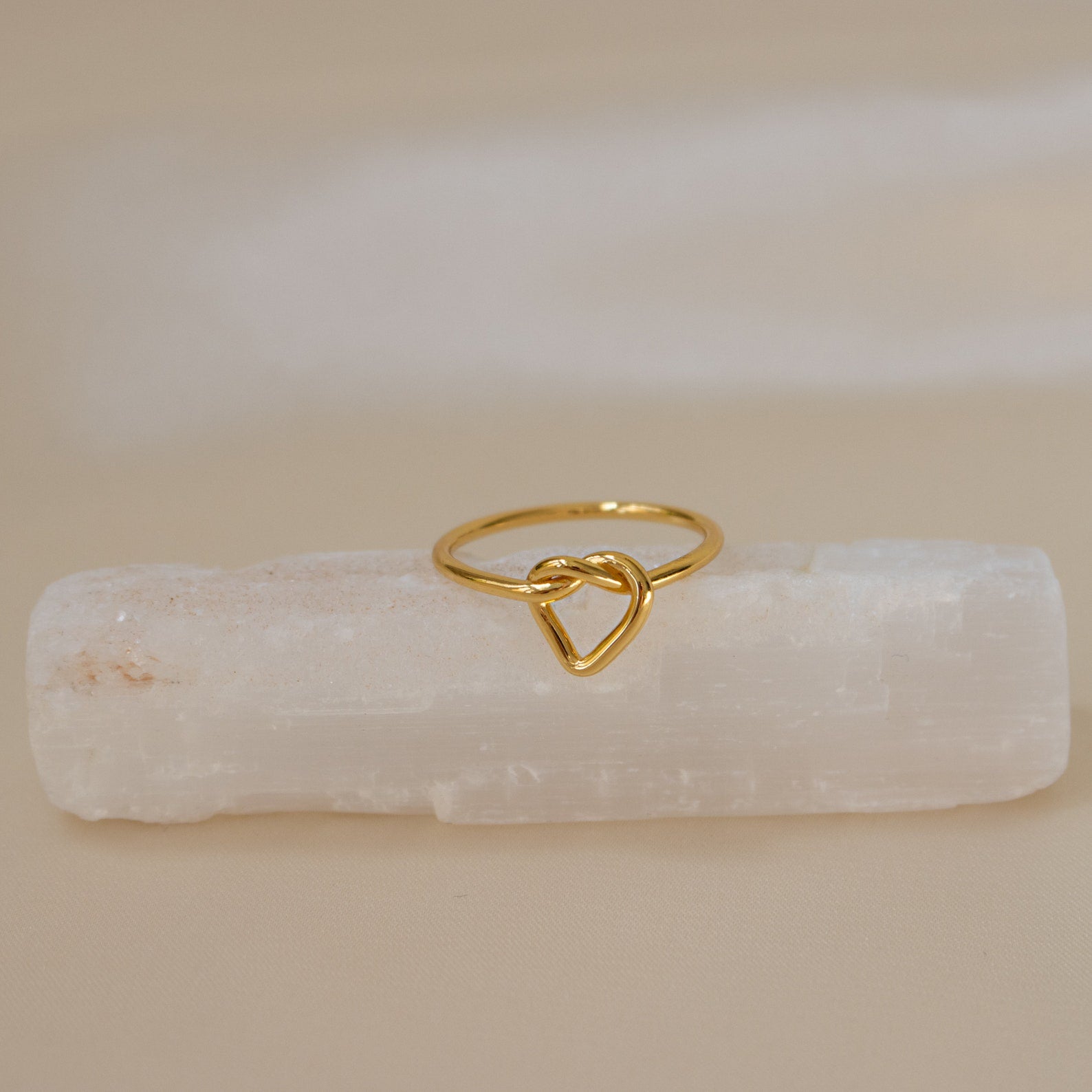 The Love Heart Knot Ring sits gracefully on a white rectangular crystal, making it an ideal promise ring or a timeless symbol of love.