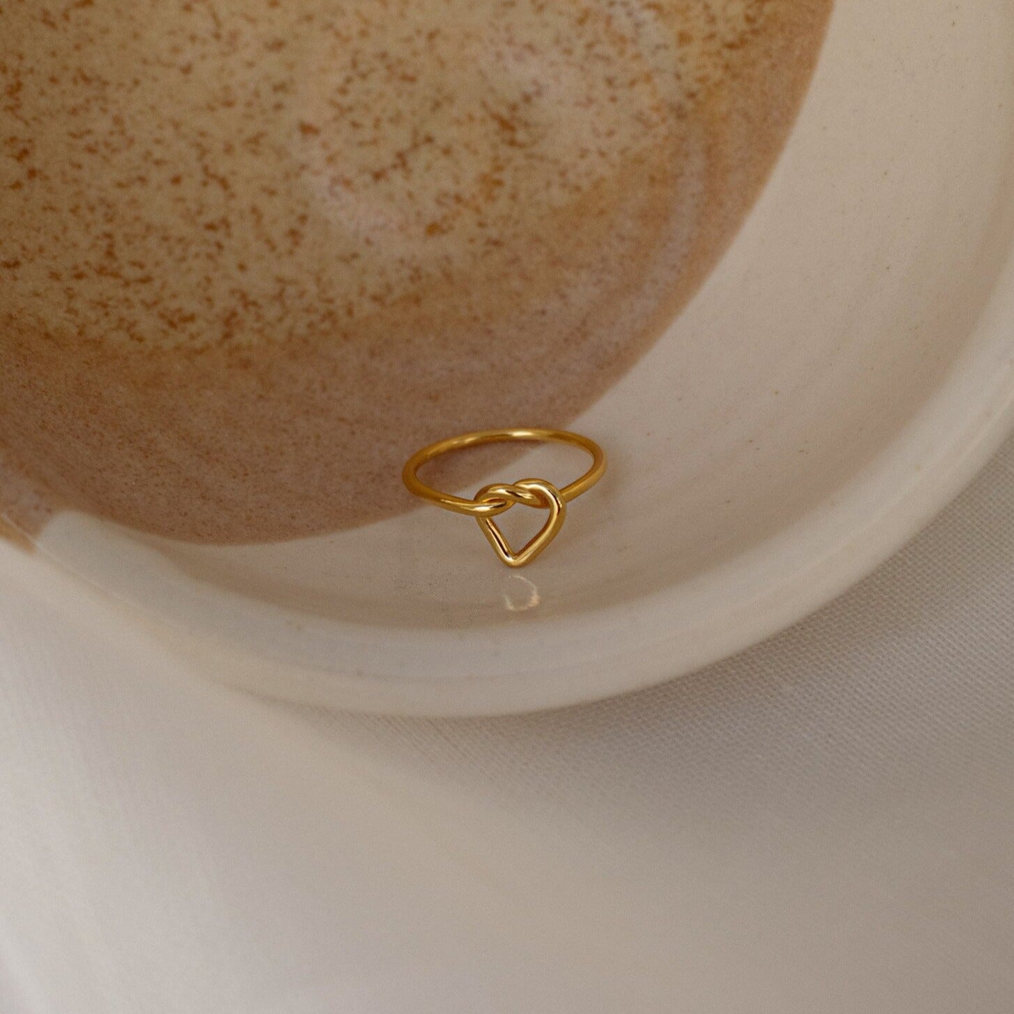 The Love Heart Knot Ring, featuring a heart-shaped knot design, is displayed gracefully in a speckled ceramic bowl.