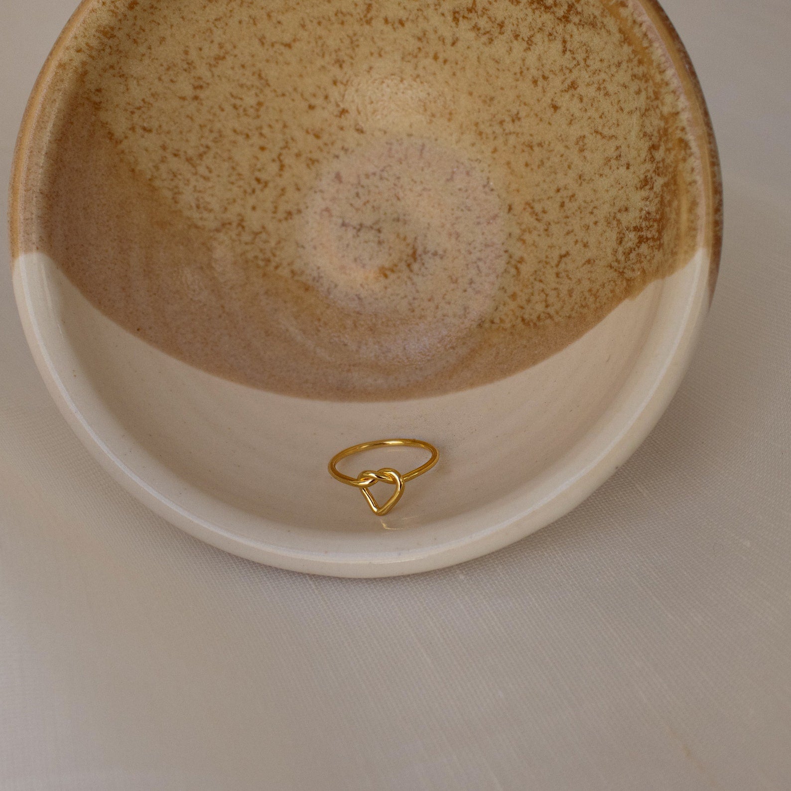 A Love Heart Knot Ring sits in a beige, speckled ceramic bowl on a light textured surface.