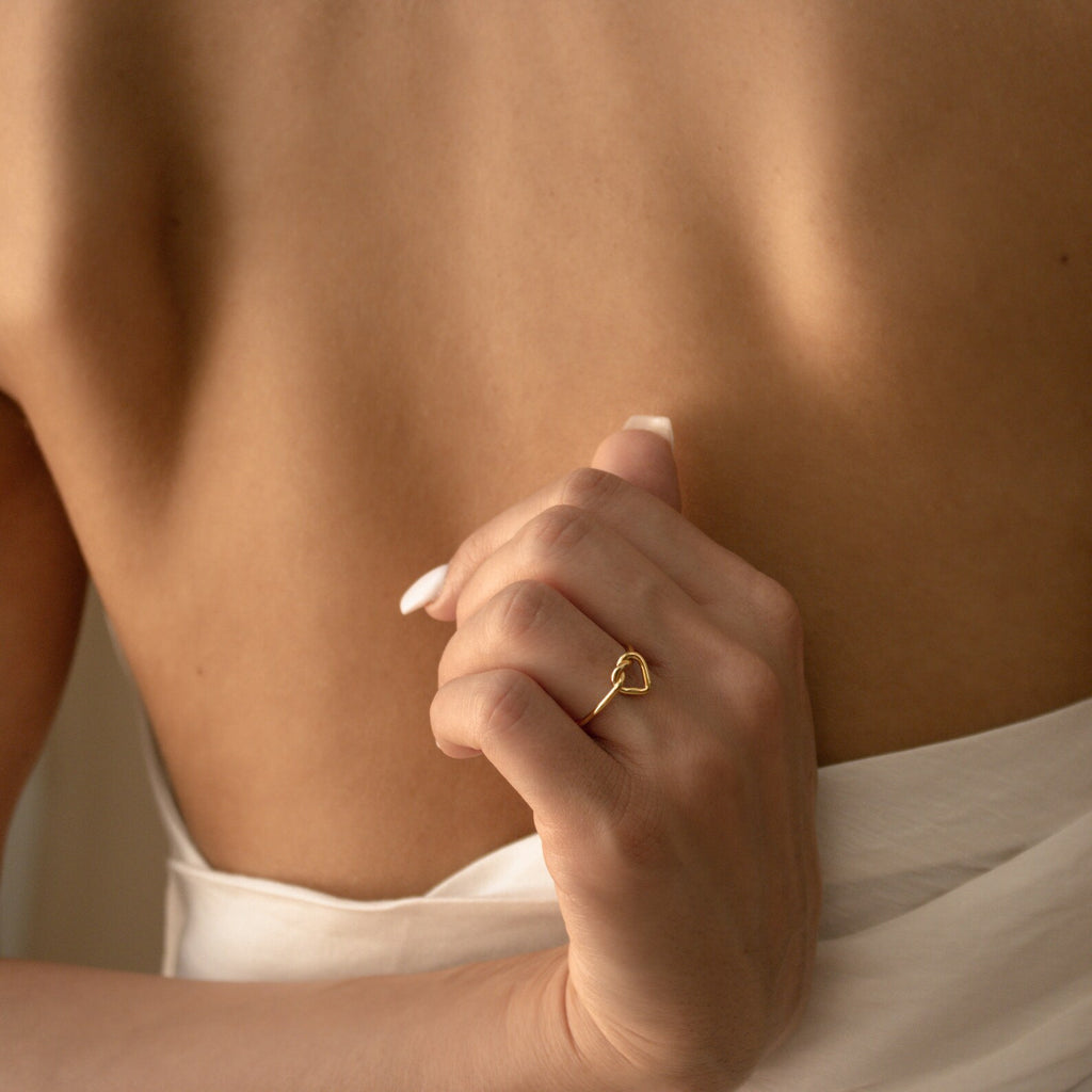 A woman with a bare back holds white fabric, wearing the Love Heart Knot Ring—a delicate golden promise ring shaped like a heart on her finger.