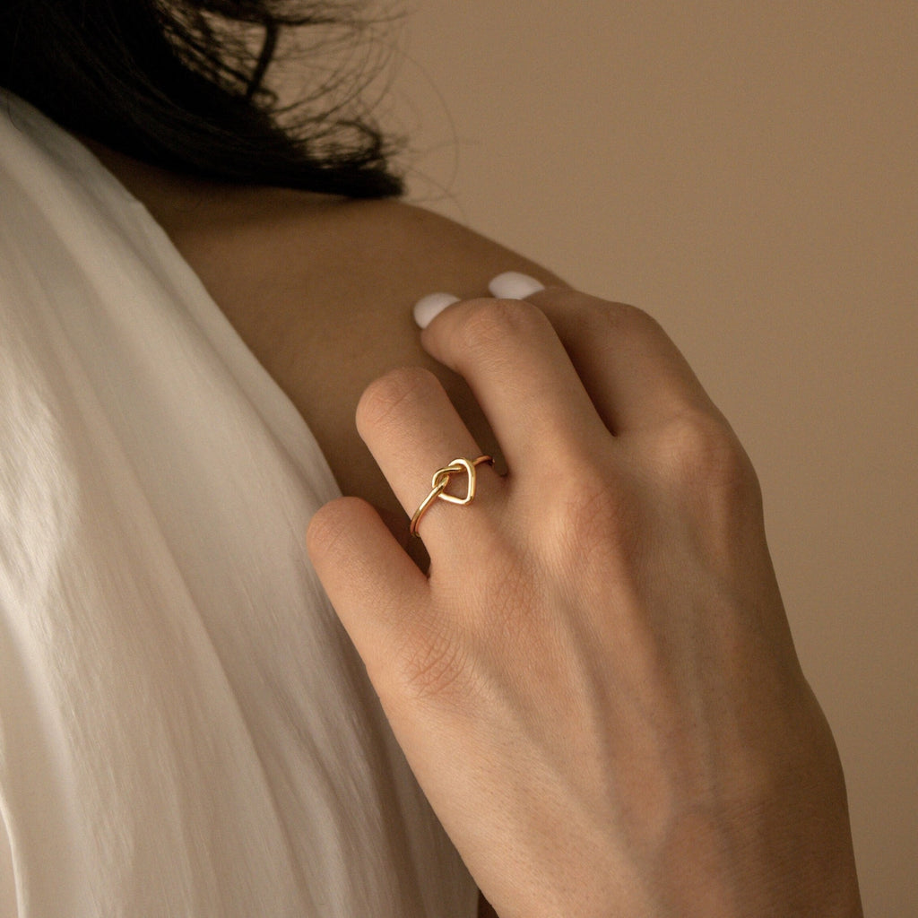 A hand wearing the delicate Love Heart Knot Ring gently touches a bare shoulder, with soft white fabric draped close by.