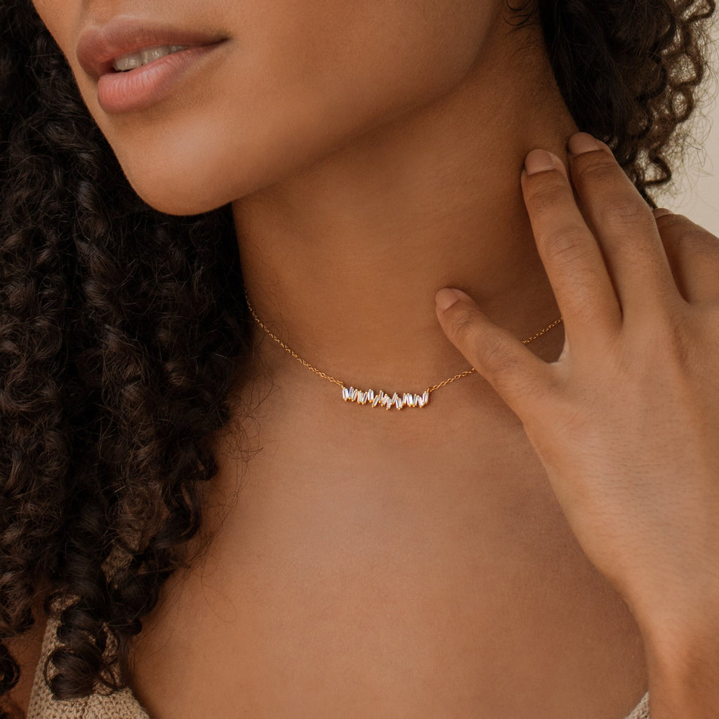 A woman with curly hair gently touches her neck while wearing the Bethany Baguette Bar Necklace, featuring a gold chain with a small, jagged bar pendant.