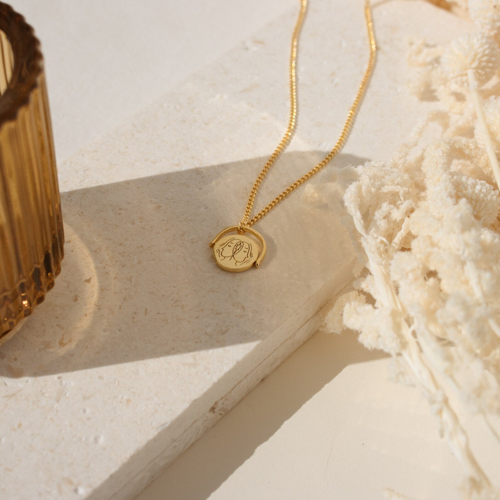 The Zodiac Spinner Necklace with an engraved astrological pendant rests on a stone surface next to a glass and dried white flowers.