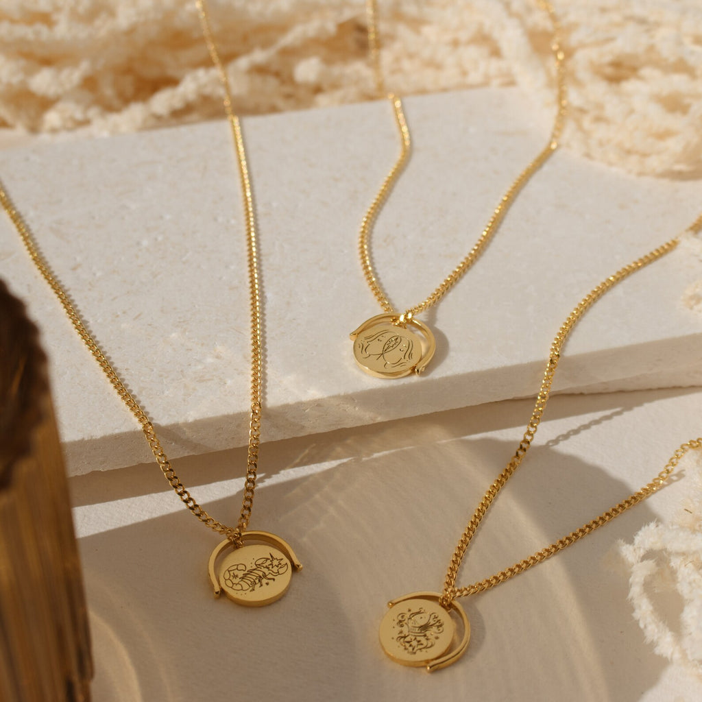 Three Zodiac Spinner Necklaces with round engraved astrological pendants are displayed on textured cream surfaces in sunlight.