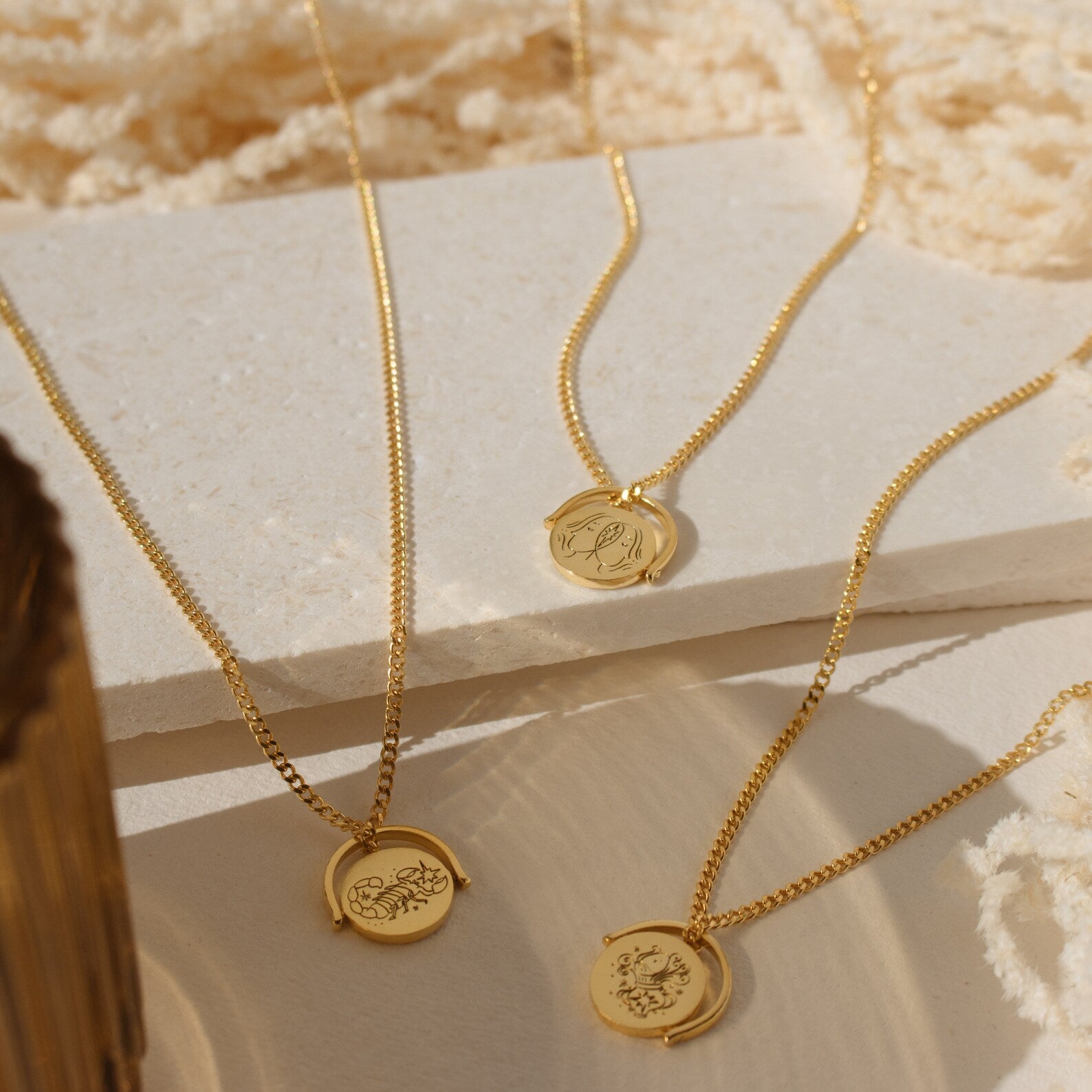 Three Zodiac Spinner Necklaces with round engraved astrological pendants are displayed on textured cream surfaces in sunlight.