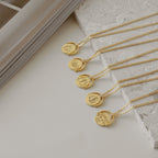 Five Zodiac Spinner Necklaces with astrological designs and personalized engraving are displayed on a white stone surface next to a book.